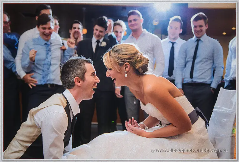 Lauren leaning from her chair laughing nose to nose with a kneeling groomsman, blokes cheering behind