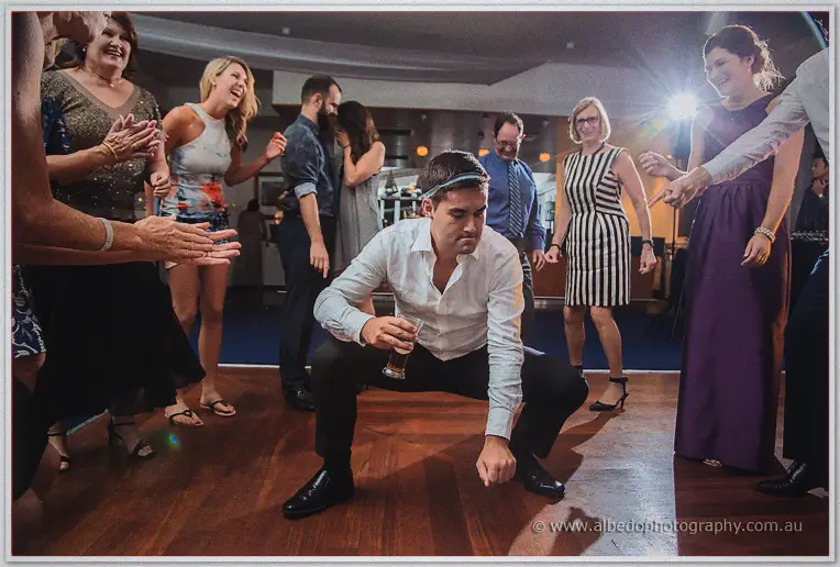 Guest in headband and beer dropping low to the floor in a squat while dancers cheer around him