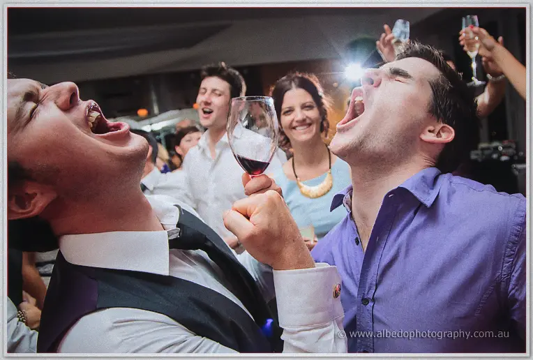 Two mates howling back to back with raised wine glass while revellers cheer at the late-night party