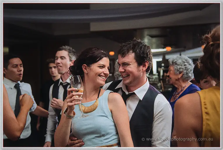 Couple in pale blue and vest sharing a grin, champagne in hand, amid the packed dim dance floor