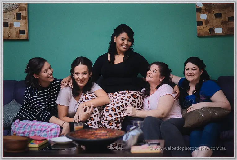 The bride laughs on a lounge with four friends in pyjamas against a teal wall before the ceremony