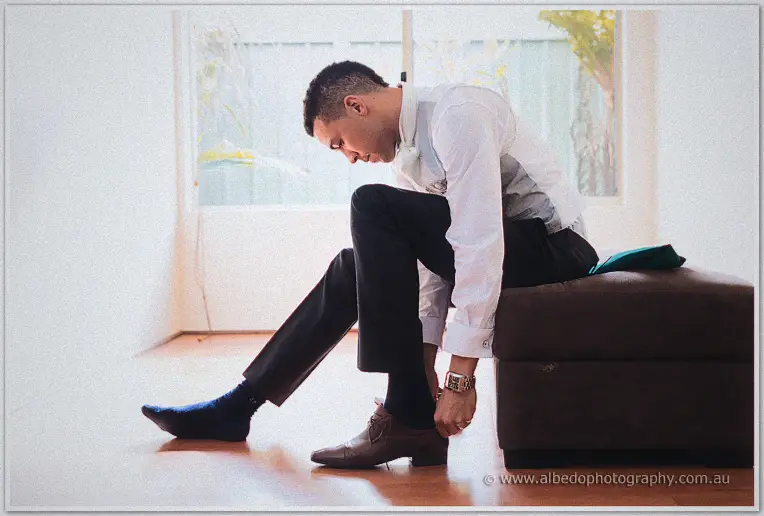 Troy hunches on an ottoman fastening a brown lace-up dress shoe beside a bright window