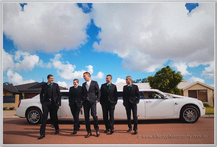 Troy and his groomsmen laugh beside a white stretch limousine under a big cloudy suburban sky