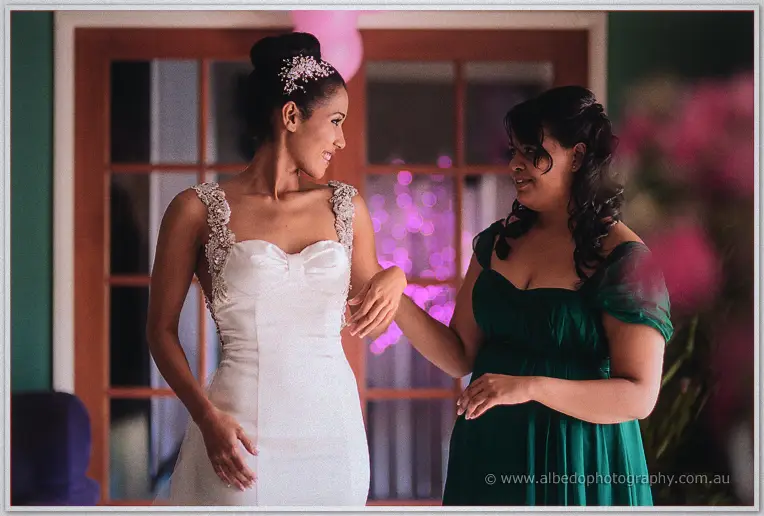 A bridesmaid in emerald adjusts the bride's dress before purple bokeh lights and timber doors