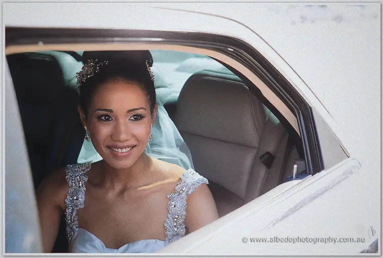 Bride Cristal smiles from a white limousine, veil and crystal headpiece catching the daylight.