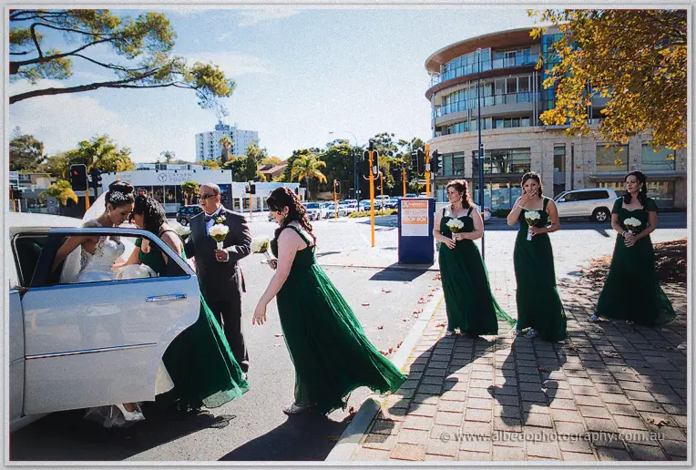 Bridesmaids in long green gowns gather on a suburban footpath as the bride steps from a stretch limousine.