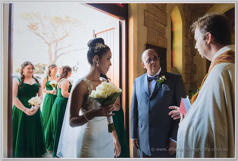 Bride with a white rose bouquet is greeted at the church door by her father in a grey suit and a robed priest.