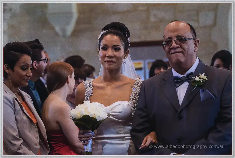 Teary bride walks arm in arm with her father down the church aisle, holding white roses.