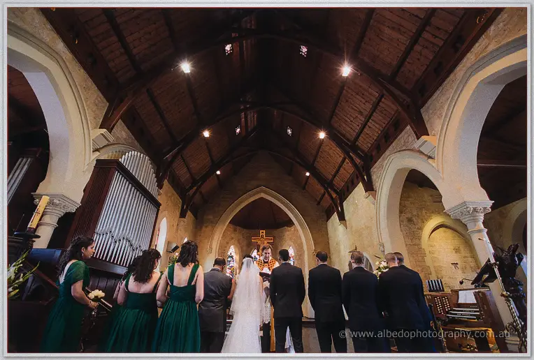 Wide view of the ceremony beneath the timber vaulted ceiling of the stone church, party facing the altar.