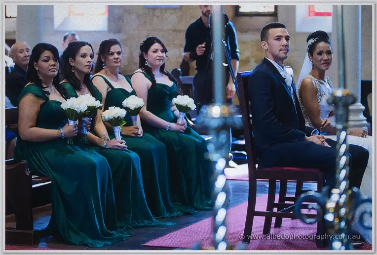 Bridesmaids in emerald gowns sit with white bouquets as the groom and bride wait near the candle stand.
