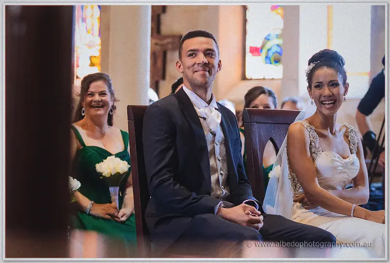 Groom Troy grins in a dark suit beside his laughing bride, green-gowned bridesmaids behind them in the church.