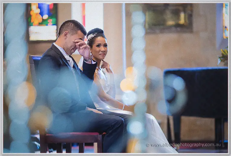 Troy wipes away a tear beside his serene bride, shot through soft candlelight bokeh inside the church.