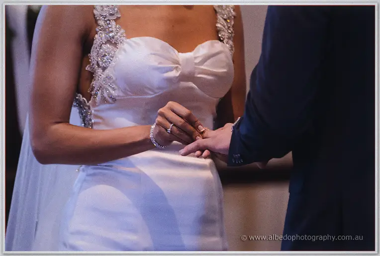 Close-up of the bride sliding a wedding ring onto Troy's finger, beaded bodice and bracelet in focus.