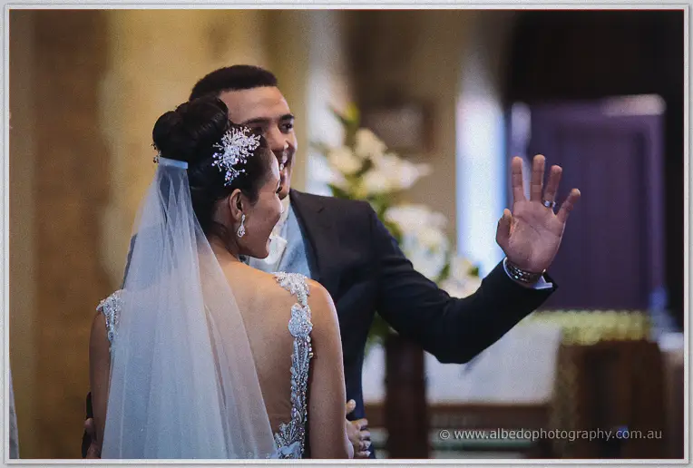 Newlyweds greet guests with a raised hand and a laugh as they turn from the altar, lace detail in view.