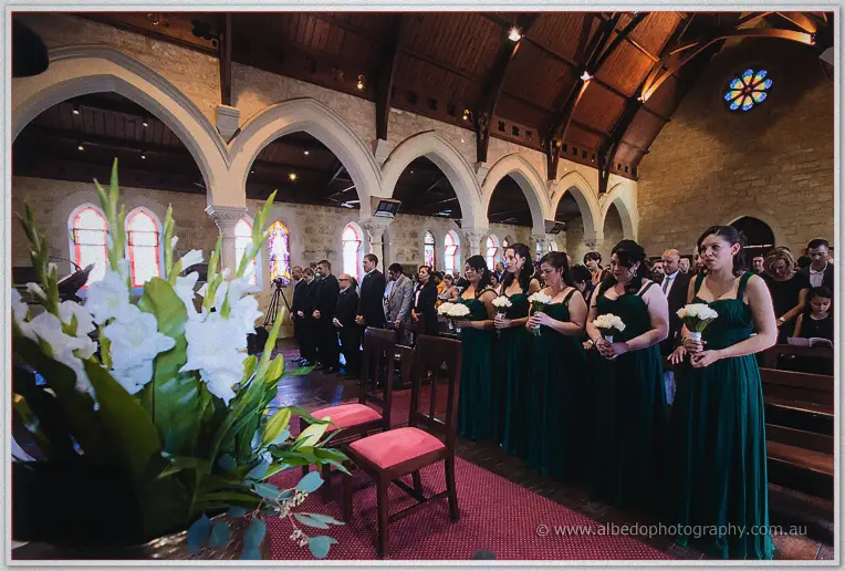 Bridesmaids in dark green hold white bouquets along the church aisle beneath the stone arches.