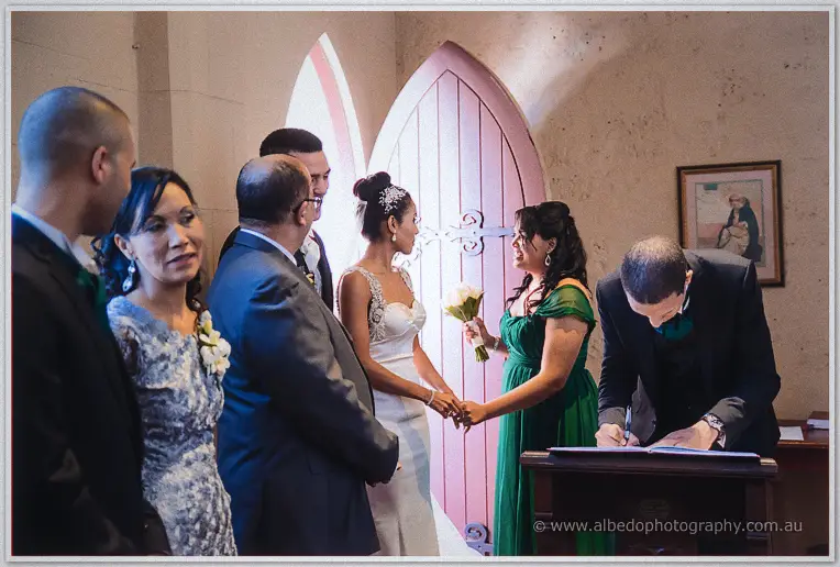 A witness signs the register at the lectern while the couple and family wait by the pink chapel door.