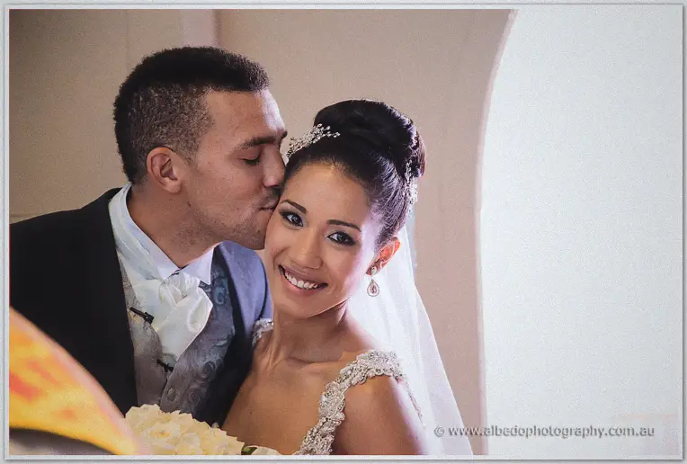 Troy kisses his bride's temple as she beams at the camera in soft window light after the ceremony.