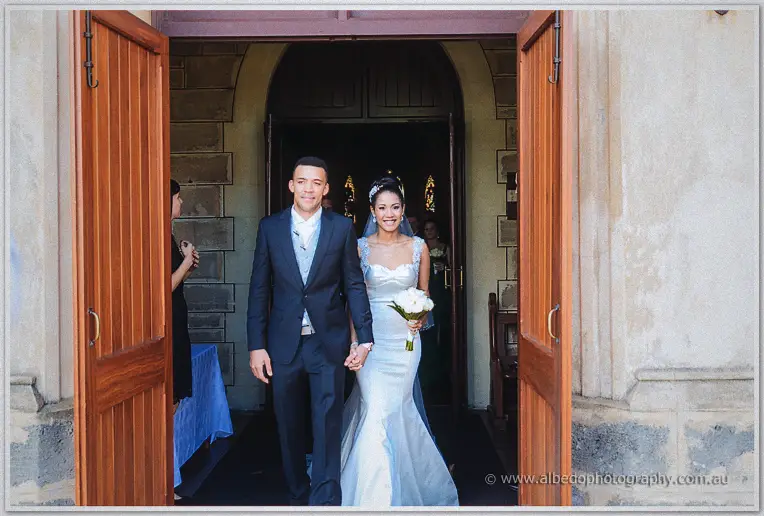 Smiling newlyweds walk hand in hand out of the timber church doors, white bouquet in hand.