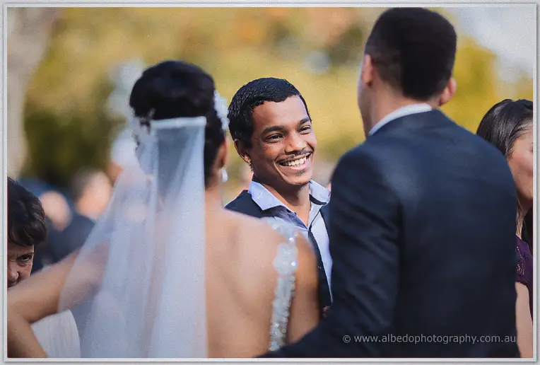A grinning guest in a navy blazer greets the veiled bride amid golden autumn light outside the church.