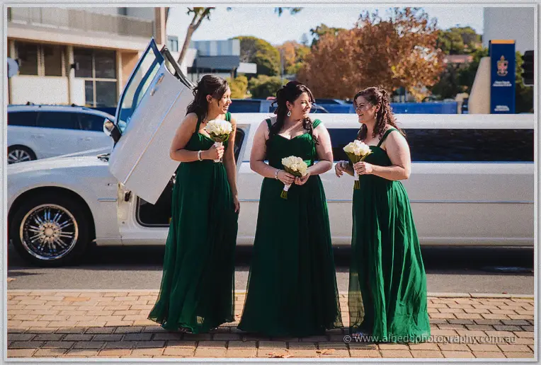 Three bridesmaids in green gowns laugh on the kerb beside the open door of the white limousine.