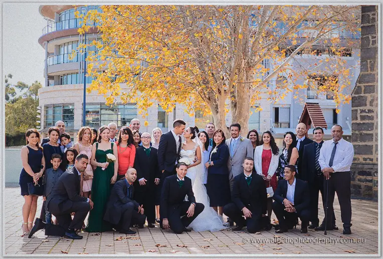 Large wedding group posed under a golden-leafed tree, the couple kissing at the centre of smiling relatives.