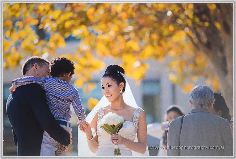 Bride holds her white bouquet while Troy lifts a small boy in a checked shirt under golden autumn trees.