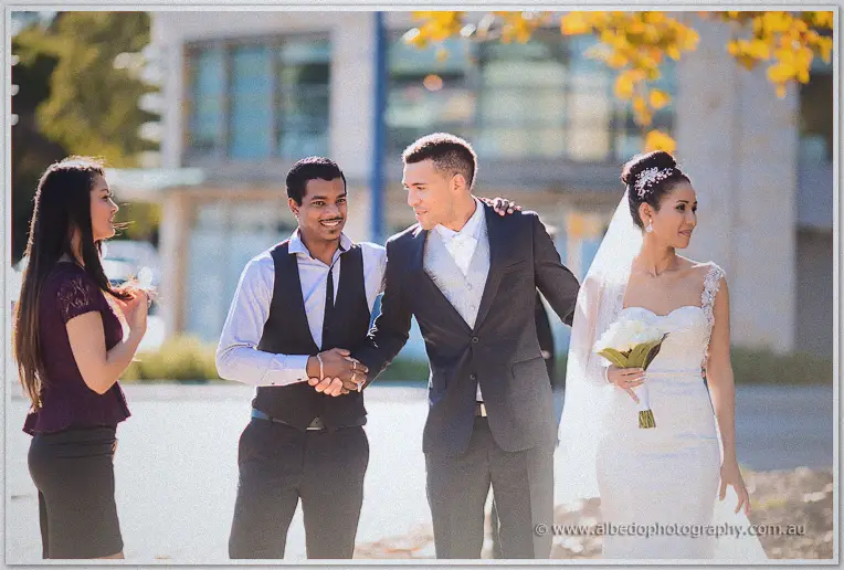 Troy shakes hands with a friend as the bride joins them on a tree-lined city street in autumn sun.