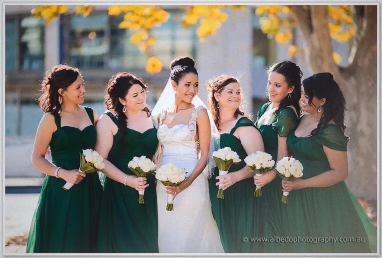 Bride in white flanked by five bridesmaids in dark green gowns holding white rose bouquets under autumn trees.