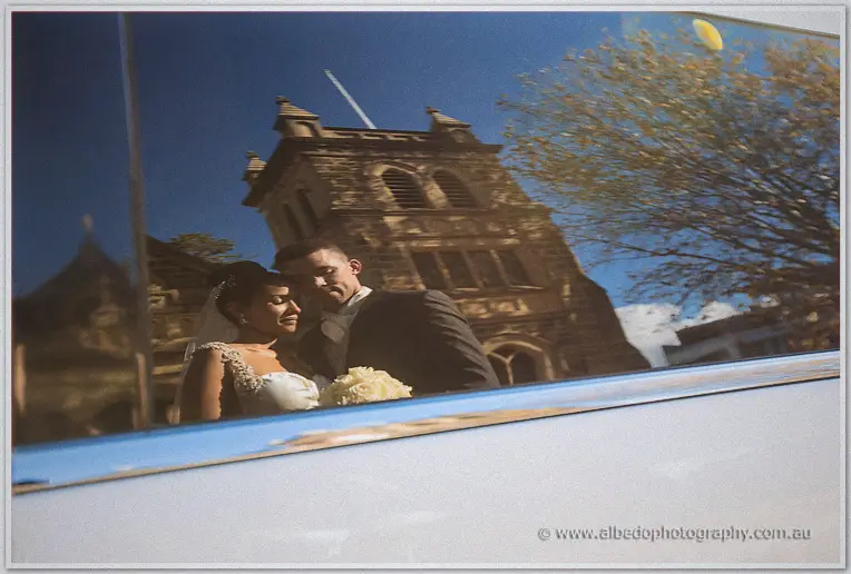 Reflection of the embracing couple and the stone church tower caught in the polished flank of the limousine.