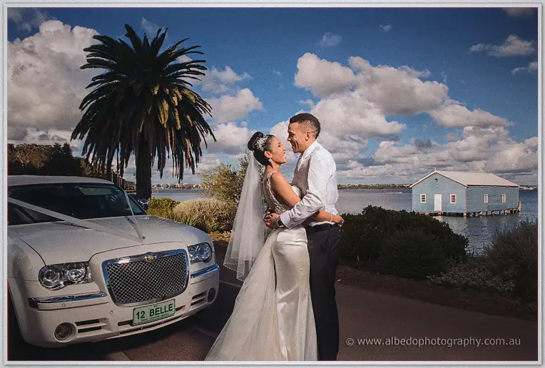 The couple embrace beside the white wedding car near the Swan River, palm and blue boathouse behind them.