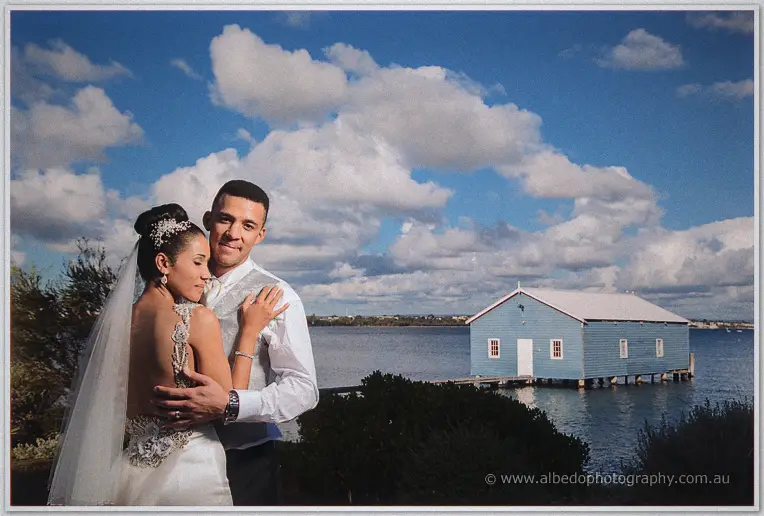 Veiled Cristal leans on Troy's chest by the river, the blue boat house floating on the calm water behind.