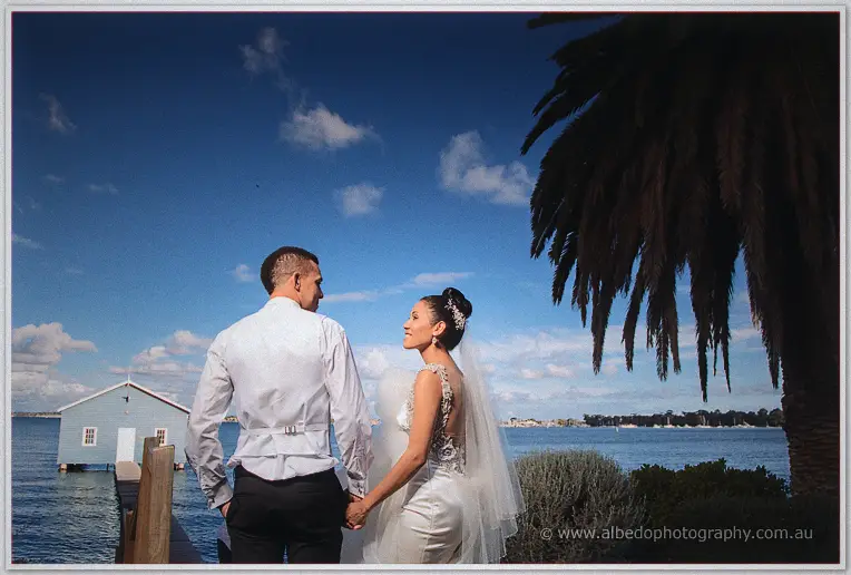 Hand in hand the newlyweds look out over the Swan River, palm fronds and the boat house framing them.