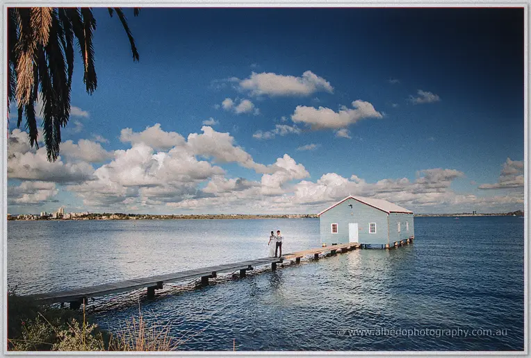 Wide scene of the couple on the long timber jetty reaching out to the blue boat house on the Swan River.
