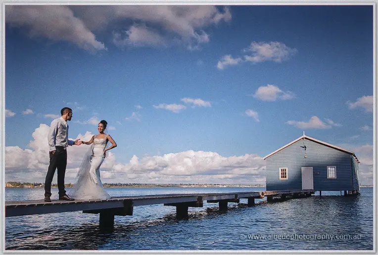 Troy holds Cristal's hand as she twirls her train on the jetty, the weathered blue boat house nearby.