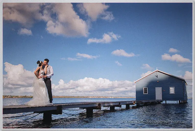 Cristal and Troy embrace on a timber jetty beneath drifting clouds beside the blue river boatshed.