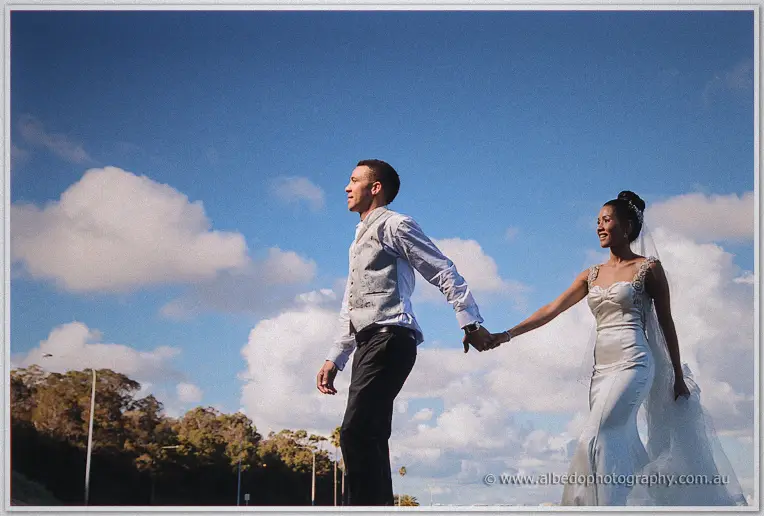 Low-angle candid of the couple walking hand in hand under a bright blue sky with drifting clouds.
