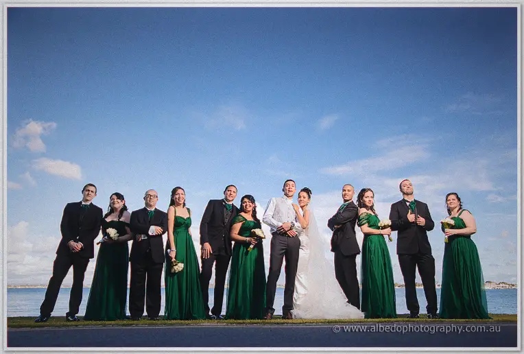 Cristal and Troy line up with their full bridal party in emerald gowns and dark suits by the water.