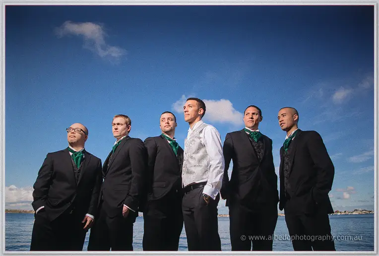 Troy and his groomsmen stand shoulder to shoulder by the water, green ties against a summer sky.