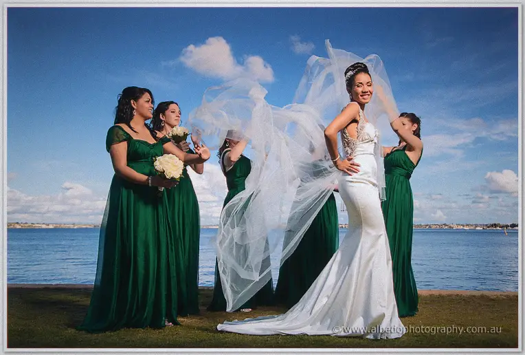 Wind lifts Cristal's veil as her bridesmaids in emerald gowns gather on the grassy foreshore.