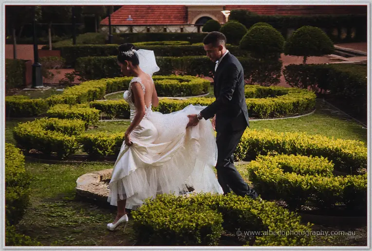 Troy gathers Cristal's train as she steps across the hedge-lined garden path at dusk.