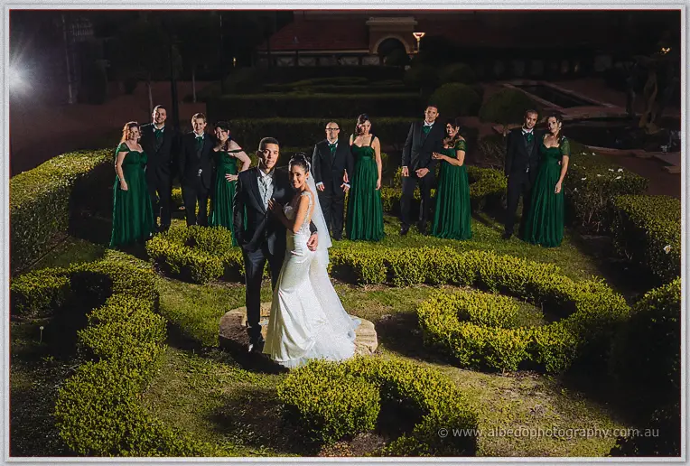 The wedding party spreads through glowing parterre hedges at night behind the embracing newlyweds.