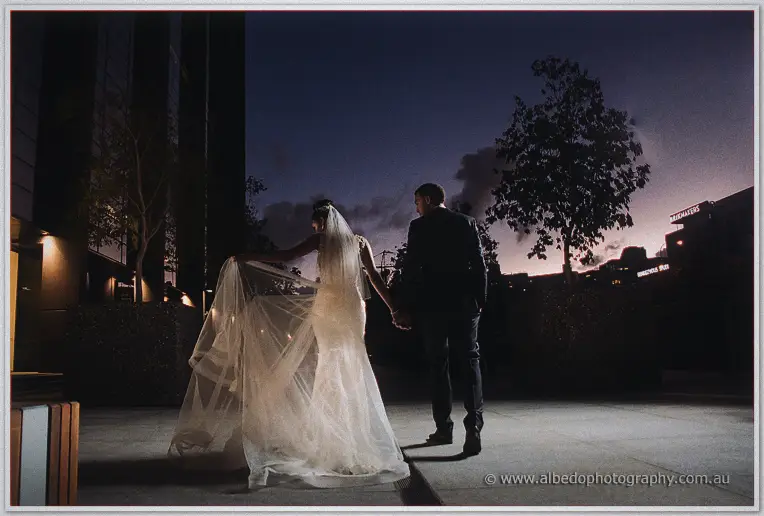 Silhouetted at deep blue dusk, Cristal lifts her billowing veil as Troy leads her through the city plaza