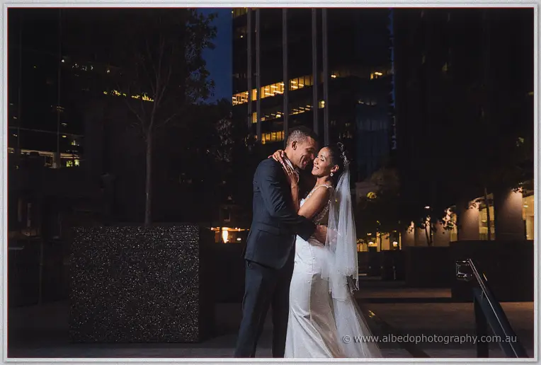 Cristal and Troy embrace in a dark plaza, warm office lights glowing in the towers behind them.
