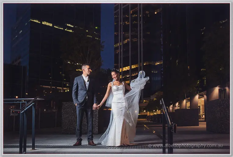 Holding hands on night steps, Cristal smiles as the breeze whips her veil toward the city towers.