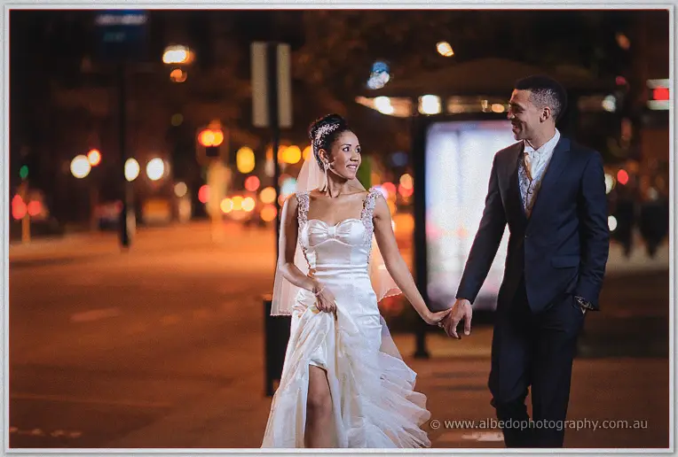 Hand in hand through warm traffic glow, Cristal gathers her gown while Troy grins across at her.