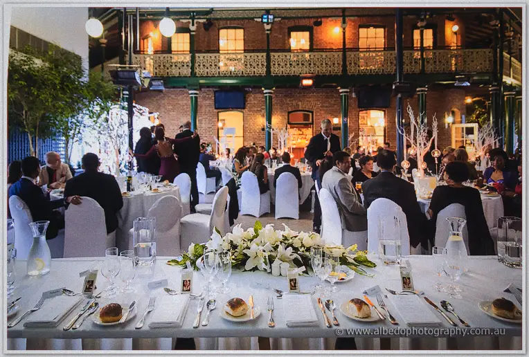 Guests fill the lamp-lit banquet hall beneath the brick balcony as dinner service begins.
