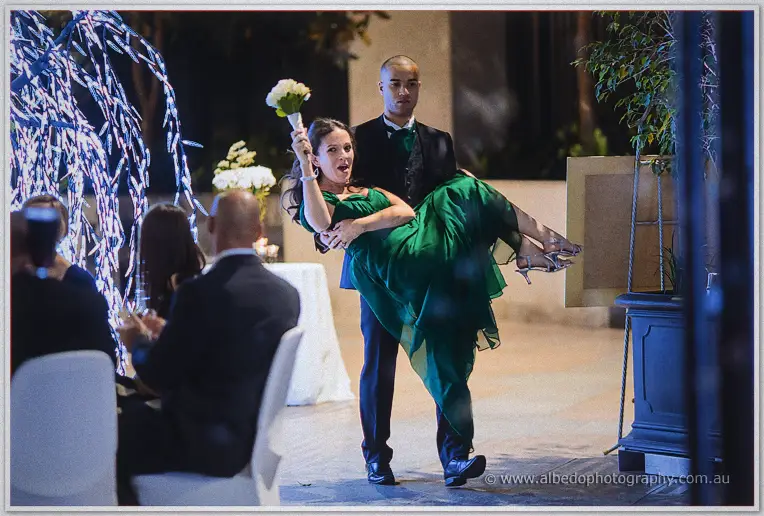 A groomsman sweeps a laughing bridesmaid in green off her feet, bouquet raised high in triumph.