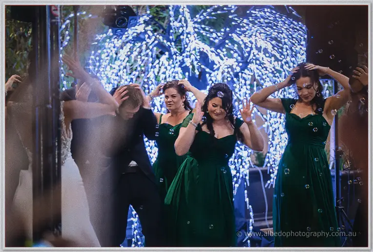 Bridesmaids in emerald duck and laugh as bubbles drift through the fairy-lit courtyard air.