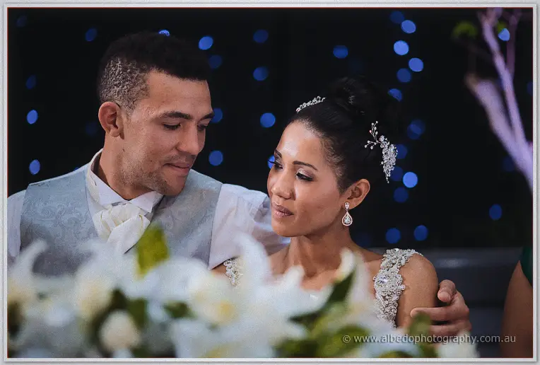 Troy's arm circles Cristal behind a bank of white lilies, faces close beneath soft blue lights.