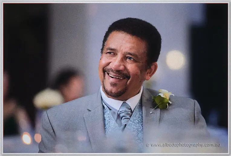 A guest in a grey suit and white rose laughs warmly during the reception speeches.